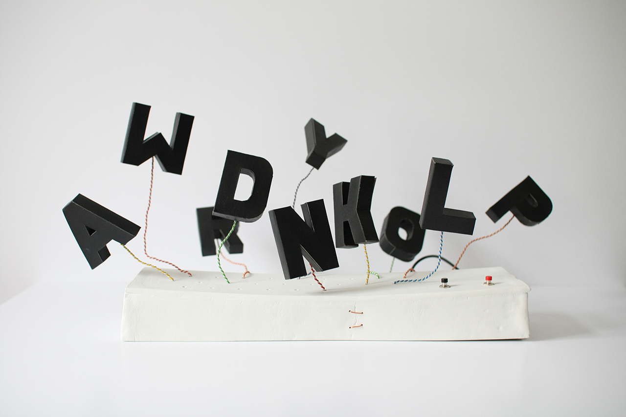 my experimental keyboard: a dozen black three-dimensional paper letters are held in the air by colourful stiff wires that emerge out a white ceramic cube